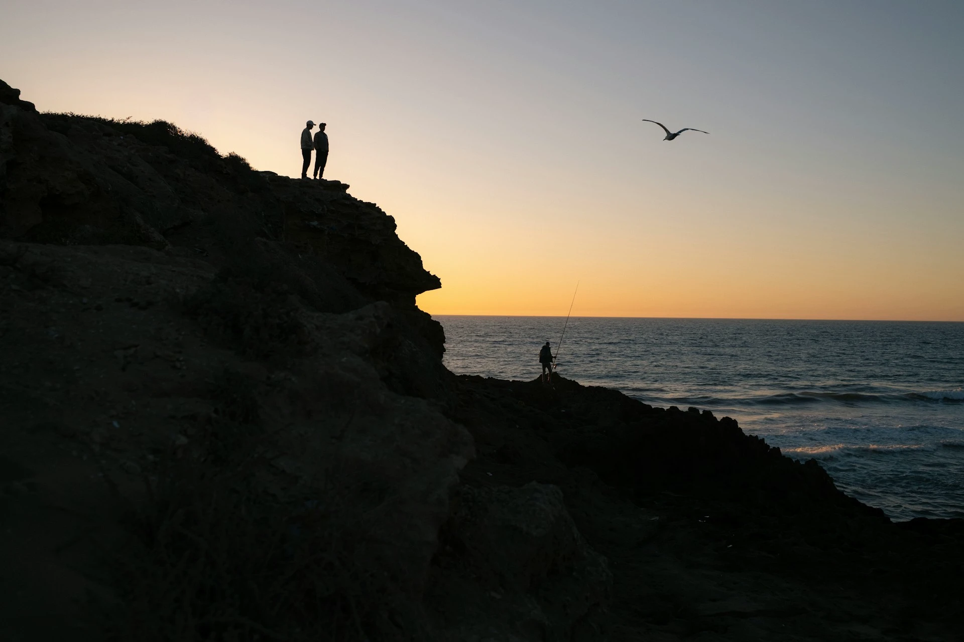 Two people look into the ocean from a short rocky cliff above while another person uses a fishing line down below.